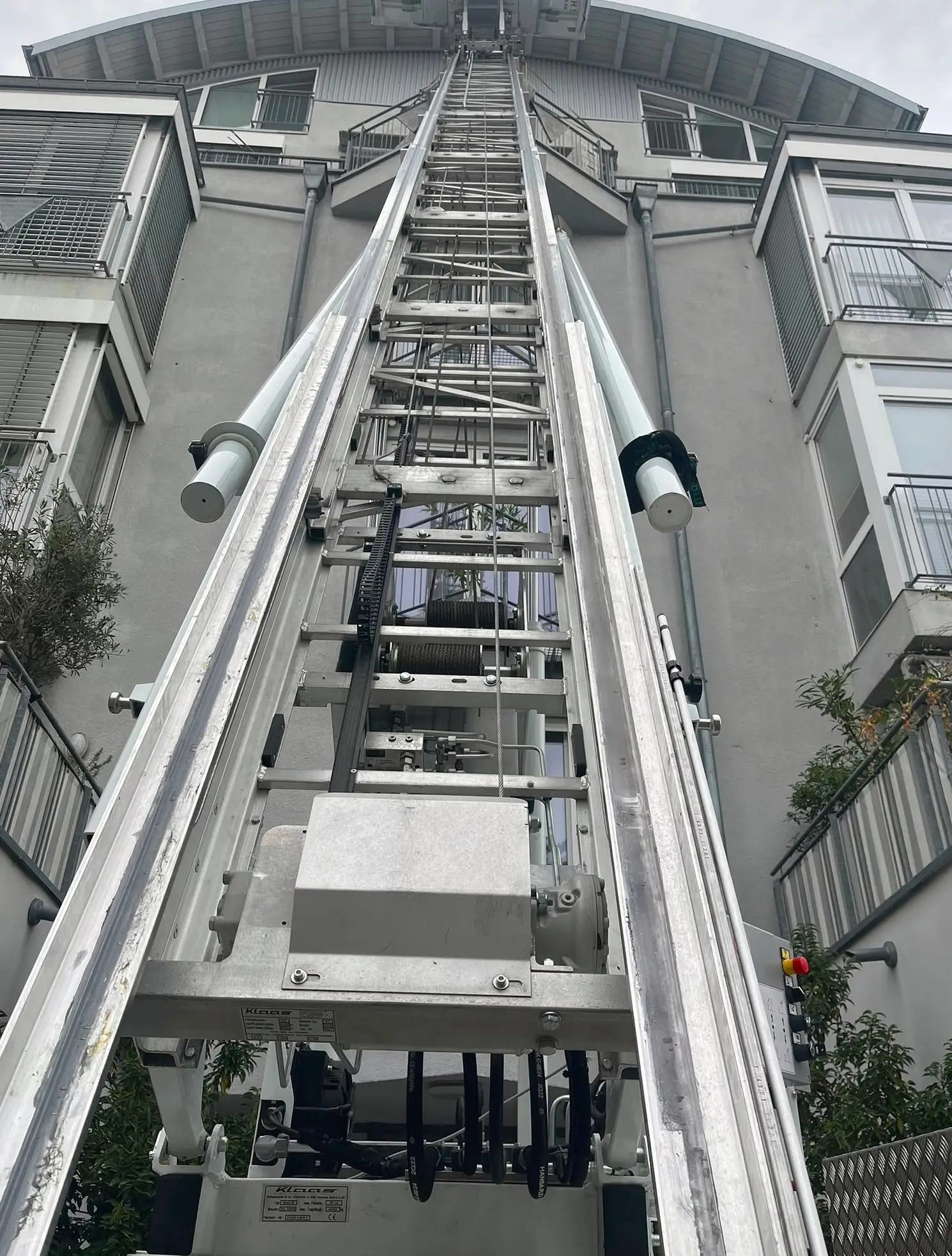 Blick von unten auf den Unterbau des Möbellifts: symmetrische Leiter, zwei Hydraulikzylinder, Seiltrommeln und ein roter Not-Aus-Schalter.