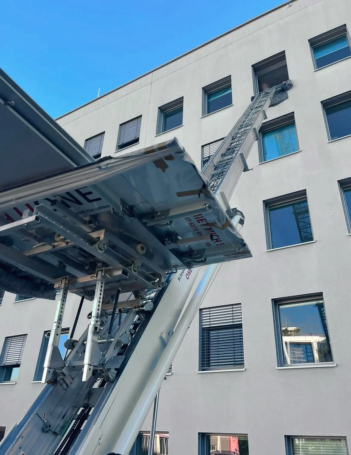 Mitarbeiter nimmt an einem offenen Fenster ein verpacktes Möbelteil von der Plattform des Möbellifts entgegen, von unten im Gegenlicht aufgenommen.
