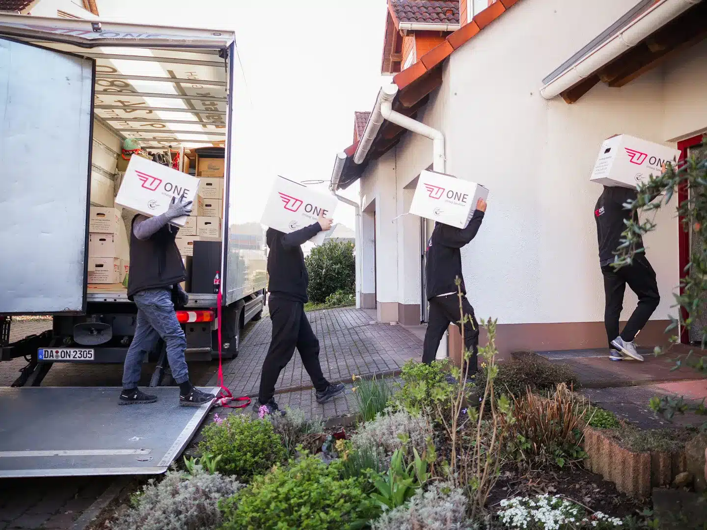 Vier Personen tragen Kisten mit der Aufschrift „EINS“ von einem Umzugswagen zu einem Haus in Darmstadt. Sie laufen unter klarem Himmel einen Weg neben dem Haus entlang. Im Vordergrund ist ein Garten zu sehen, im Hintergrund sind Dächer zu sehen – eine ideale Kulisse für einen Umzug an einem so schönen Tag.