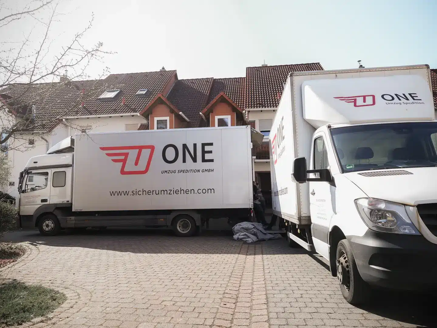 Zwei Umzugswagen mit der Aufschrift „ONE“, die die Effizienz eines reibungslosen Umzugs symbolisieren, parken in der Einfahrt eines Wohnhauses vor einem Haus mit Ziegeldach. Die weißen Wagen mit den roten Logos heben sich von der gepflasterten Umgebung ab, während links ein Ast hervorschaut.