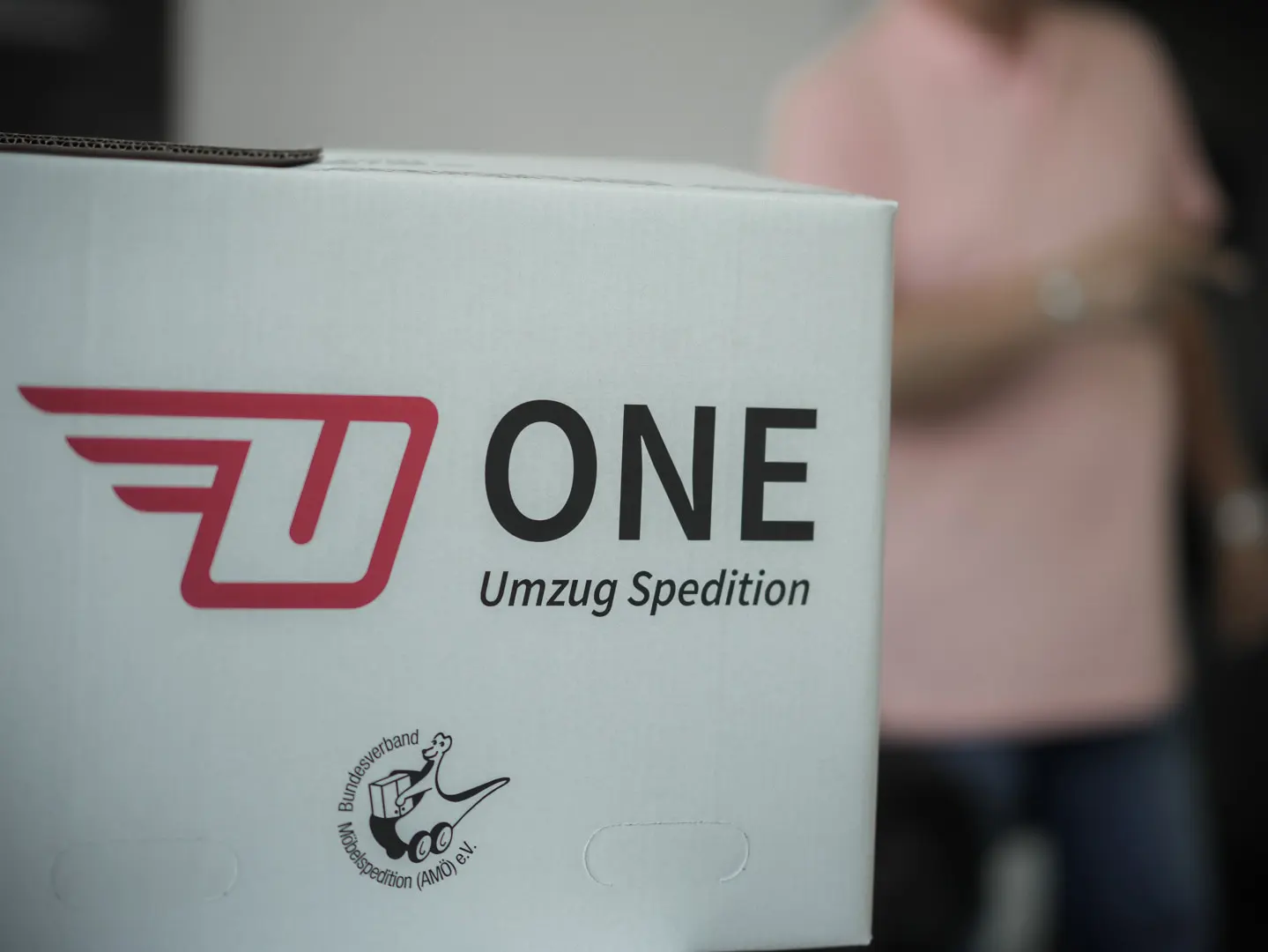 Karton mit der Aufschrift „ONE Umzug Spedition“ und dem Logo mit einem stilisierten U und Flügeln. Ein kleineres Symbol zeigt einen Schuh und einen Mann, der Umzugskartons trägt. Im Hintergrund verleiht eine verschwommene Person in einem rosa Hemd der Szene Tiefe.