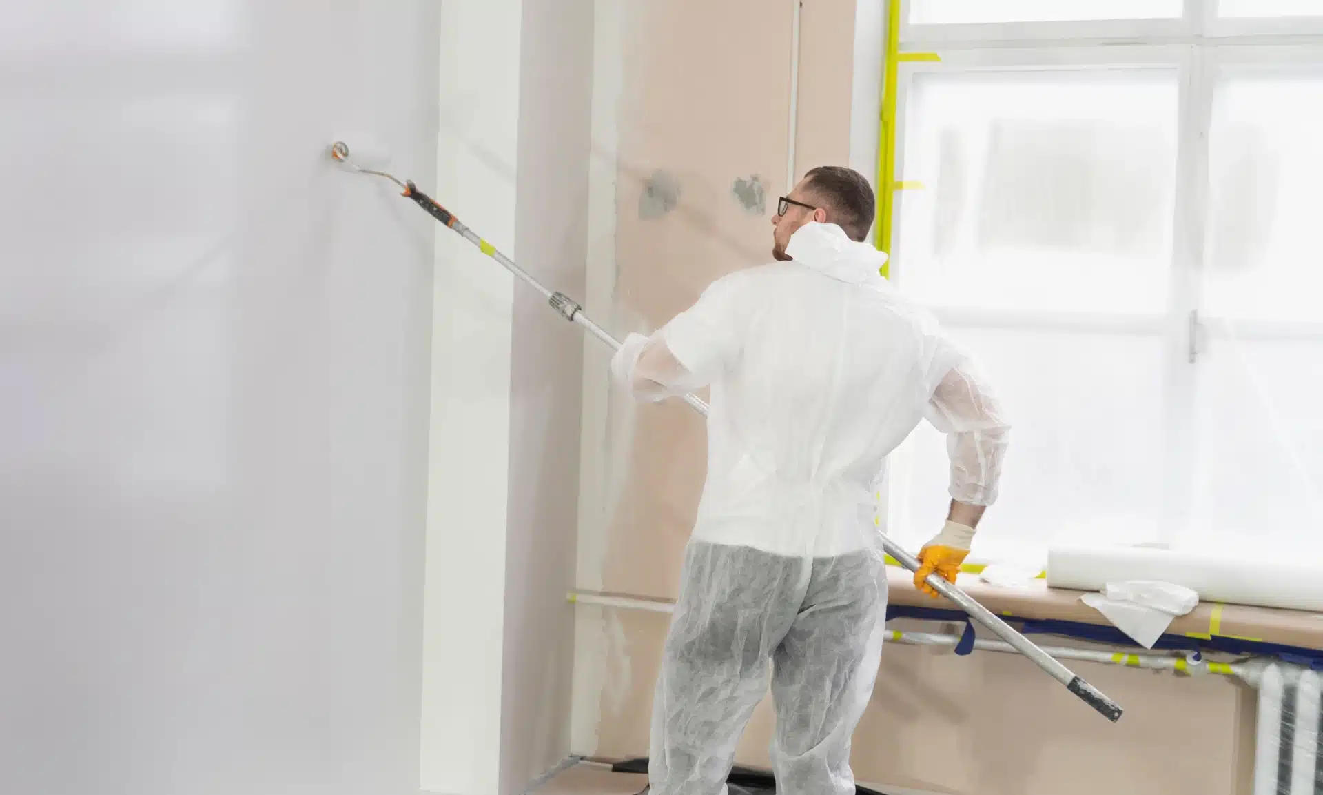 Eine Person in Schutzkleidung und Schutzbrille streicht mit einer Rolle mit langem Stiel eine Wand. Der Raum, der vermutlich gerade renoviert wird, ist teilweise mit Fenstern abgedeckt. Mit Blick auf die Wand trägt sie eine helle Farbe auf und verkörpert damit die sorgfältige Handschrift eines Malerfachmanns.