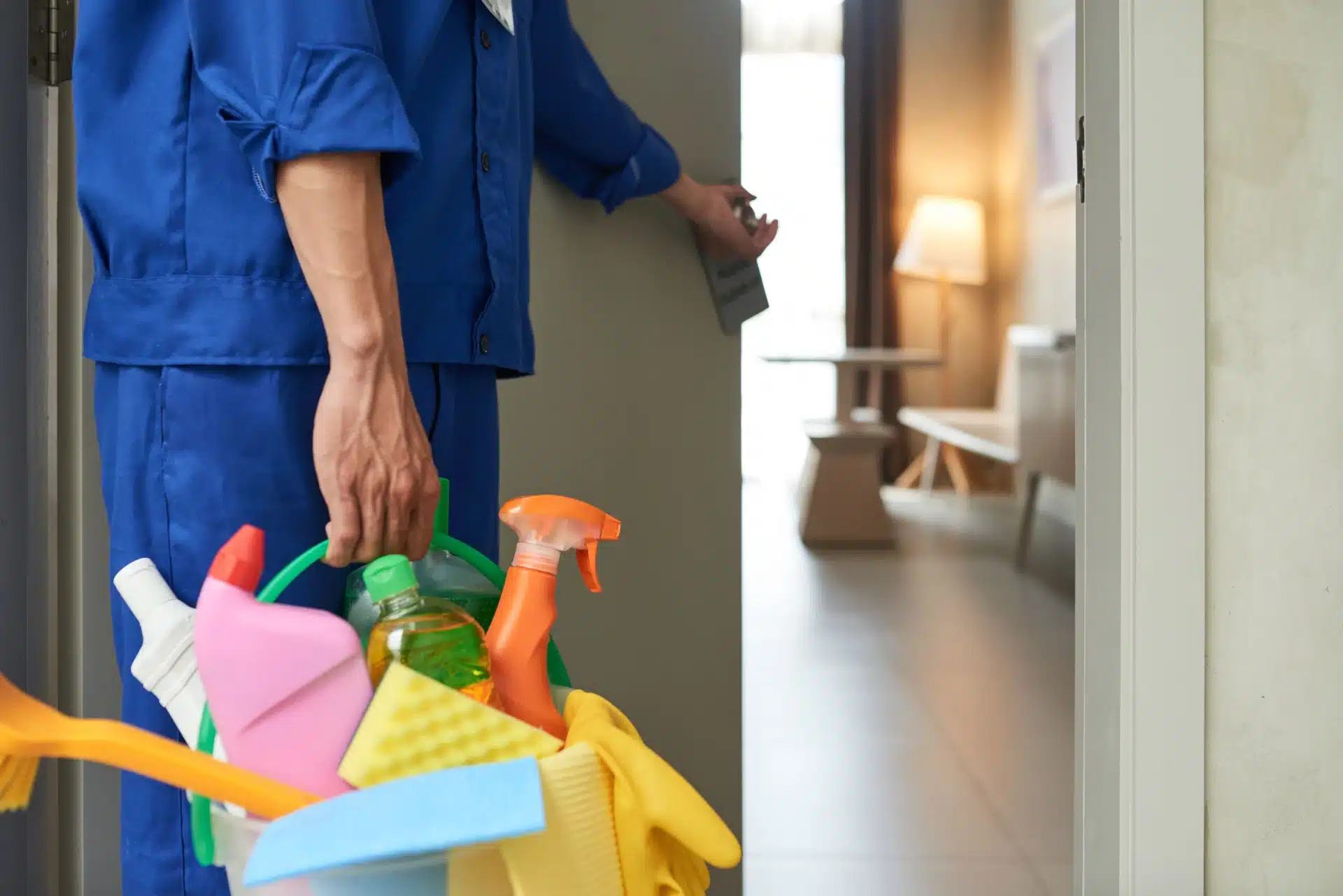 Eine Person in blauer Uniform öffnet eine Tür und hält einen Behälter mit Reinigungsmitteln in der Hand, darunter Sprühflaschen, Schwämme und eine Bürste. Der Raum ist hell erleuchtet, eine Lampe brennt, und ein Holztisch ist sichtbar – perfekt für einen Umzugsreinigungsservice, der dafür sorgt, dass alles glänzt.